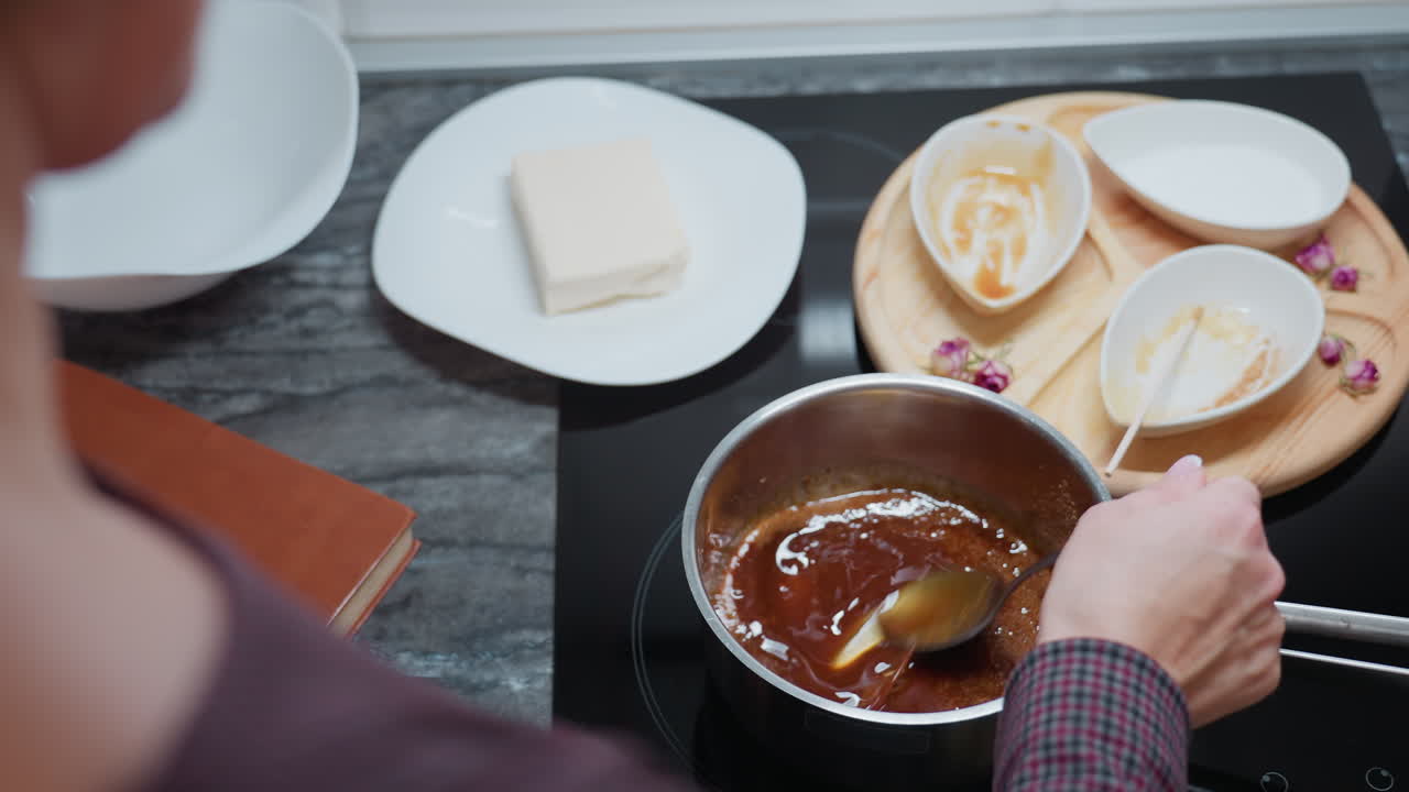 vista parcial de una persona mezclando sopa en una olla de acero inoxidable con una cuchara, ingredientes mezclándose en líquido, bandeja de madera con cuencos y utensilios usados en la encimera de la cocina de mármol oscuro