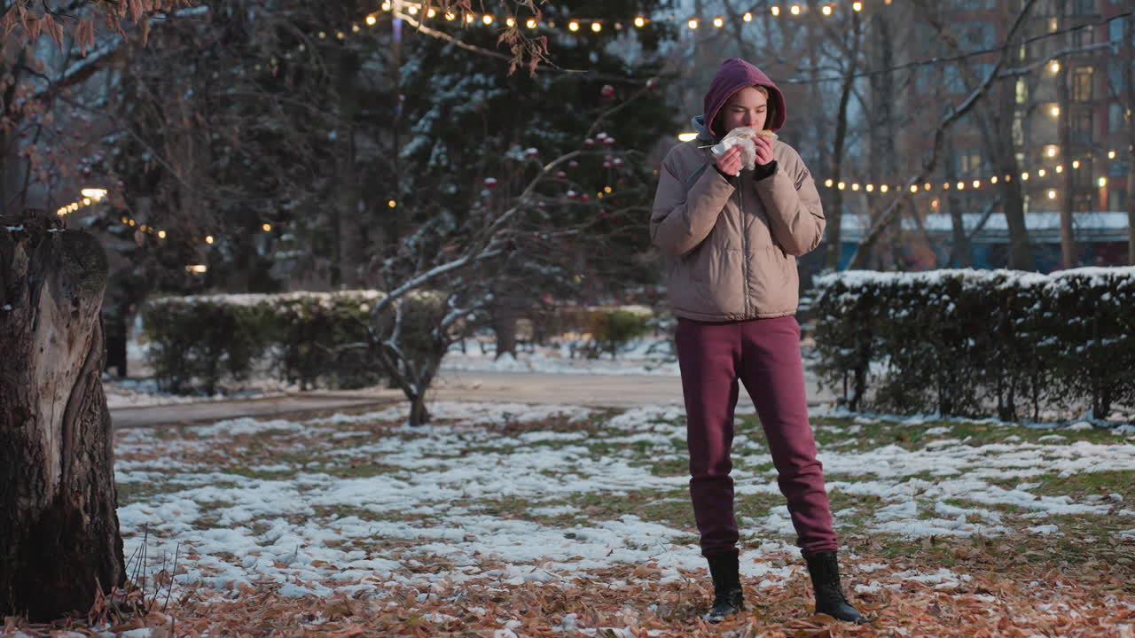 белая леди использует салфетку, чтобы очистить рот, держа в руке закуску, стоя в парке зимой с огнями боке в размытом фоне, люди ходят вокруг