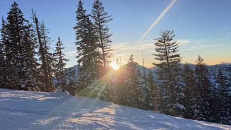 Tilt-down-winter-landscape-scene-of-a-path-on-a-ski-resort-in-the-rocky-mountains-in-Utah-with-skiers-passing-by-during-a-bright-colorful-sunset-passing-through-snow-covered-pine-trees-on-a-spring-day