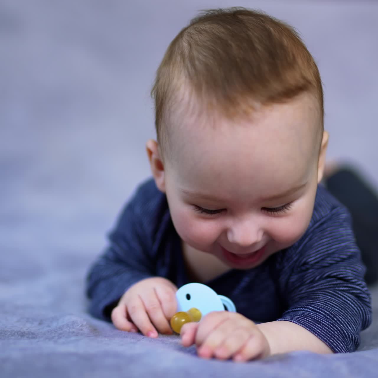 Little infant boy resting on the bed lying on belly. Adorable child holds a pacifier, looks at it and plays with it. Grey backdrop