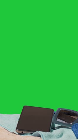 Vertical-Video-Summer-Holiday-Concept-Of-Sunglasses-Mobile-Phone-Beach-Towel-On-Sand-Against-Green-Screen