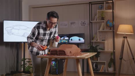hombre asiático diseñador de automóviles rociando y usando esponja para suavizar la superficie de la escultura de arcilla de automóvil en el estudio