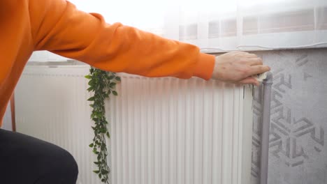 the man warms his hands on the heating radiator by the wall. cold in the apartment, poor heating system.