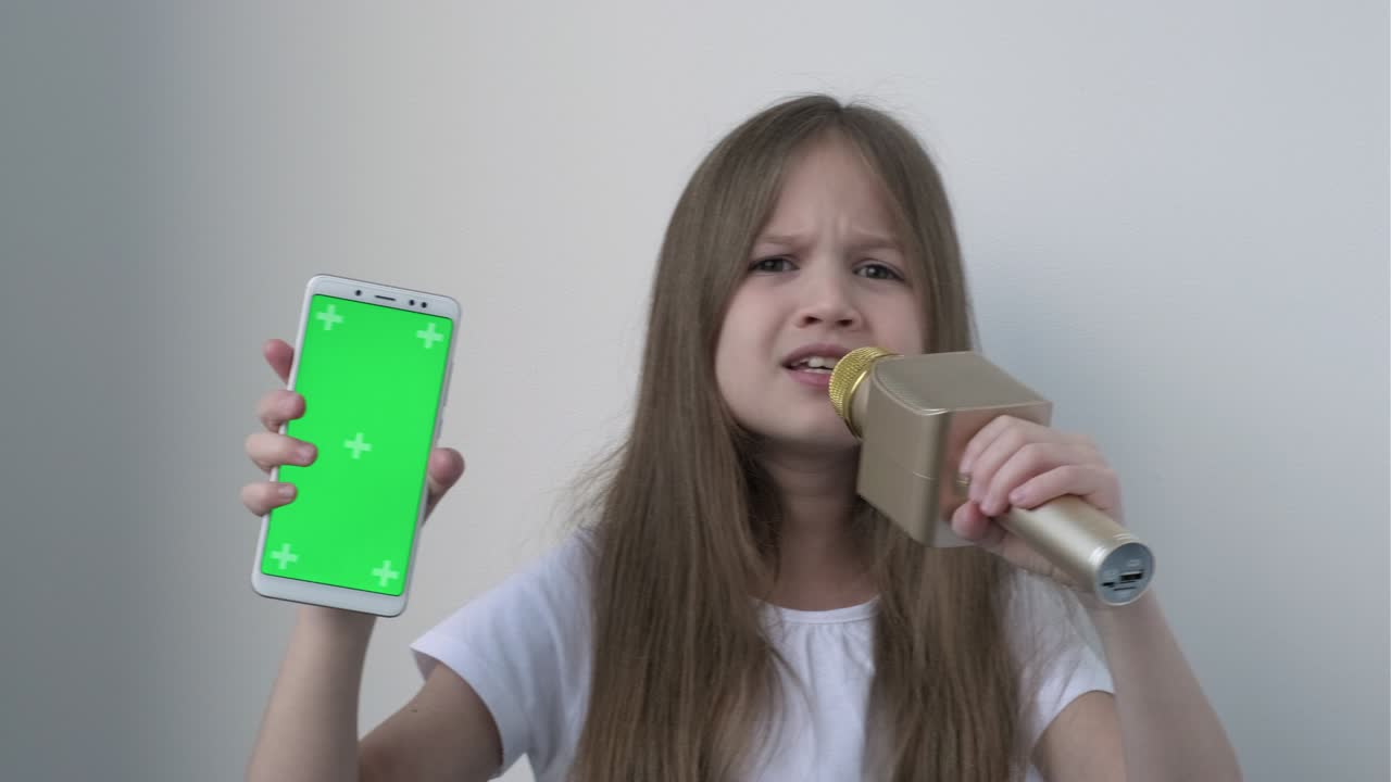 la niña muestra el teléfono con una pantalla verde y canta una canción en un micrófono de karaoke inalámbrico. chromakey es un teléfono inteligente de pantalla verde configurado para publicidad. tecnología, aparatos y niños