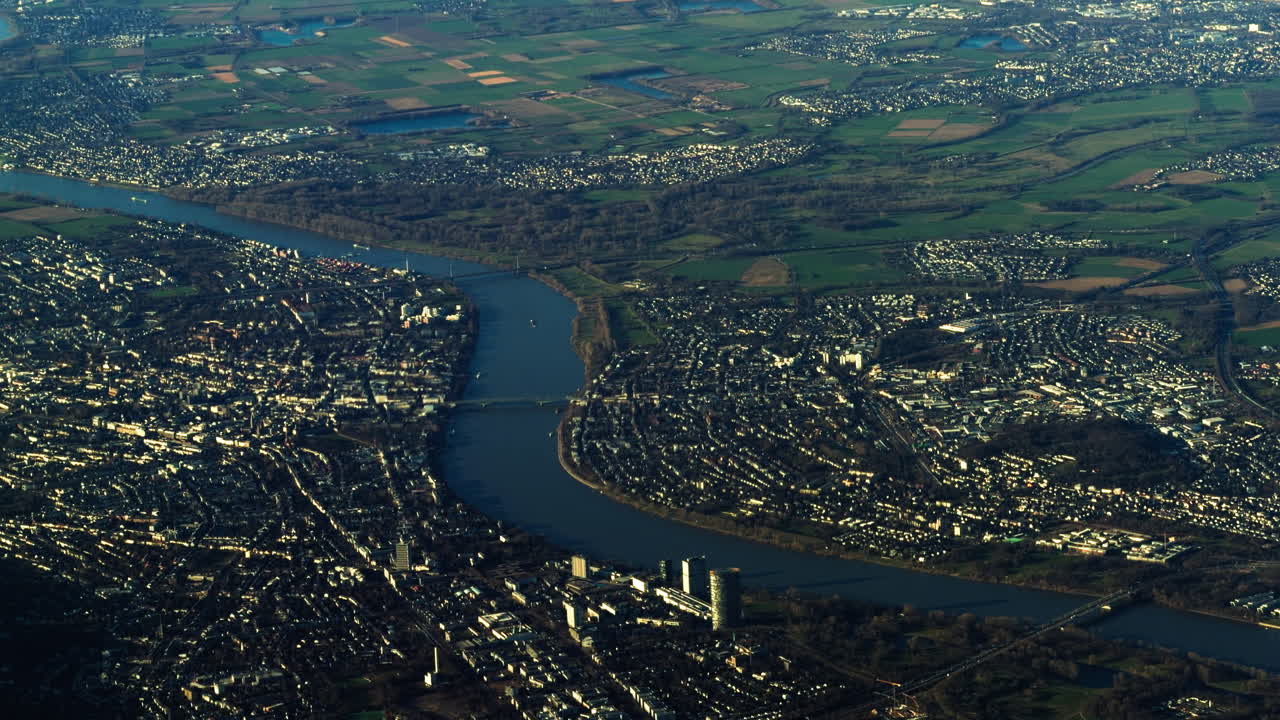 Vista aerea del fiume Reno che attraversa un paesaggio urbano vicino a Colonia in Renania Settentrionale-Vestfalia, mostrando ponti, quartieri residenziali e campi di campagna che circondano una grande città