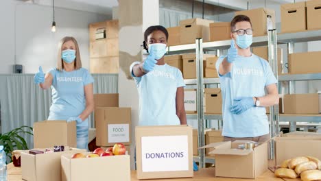 multi-ethnic group of facial mask volunteers beckoning with thumb up and looking at camera in a charity warehouse