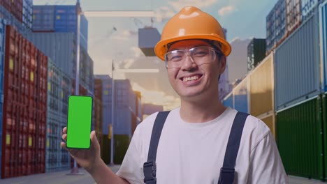 Close Up Of Asian Man Worker Wearing Goggles And Safety Helmet Smiling And Showing Green Screen Smartphone To The Camera While Standing At Container Yard Warehouse