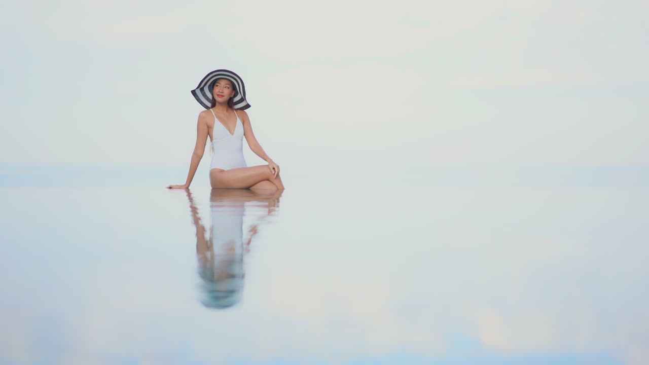 азиатская женщина в белом купальном костюме и большой черно-белой шляпе сидит на краю бесконечного бассейна и позирует