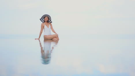 asian woman wearing white bathing suit and large black and white hat sitting on edge of infinity swimming pool posing