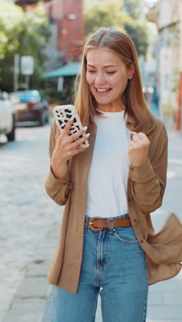 happy young woman using smartphone celebrating win good message news clenching fists on city street