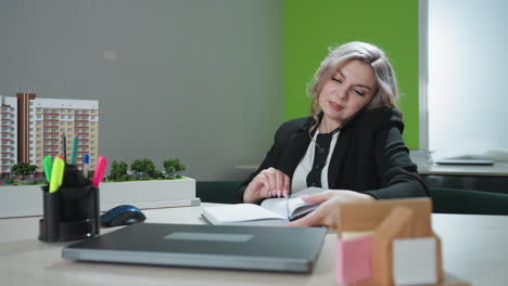 real estate sales person makes client call while seated in office, opens book and starts jotting notes, surrounded by architectural model, laptop, pens, and desk plant in brightly lit workspace