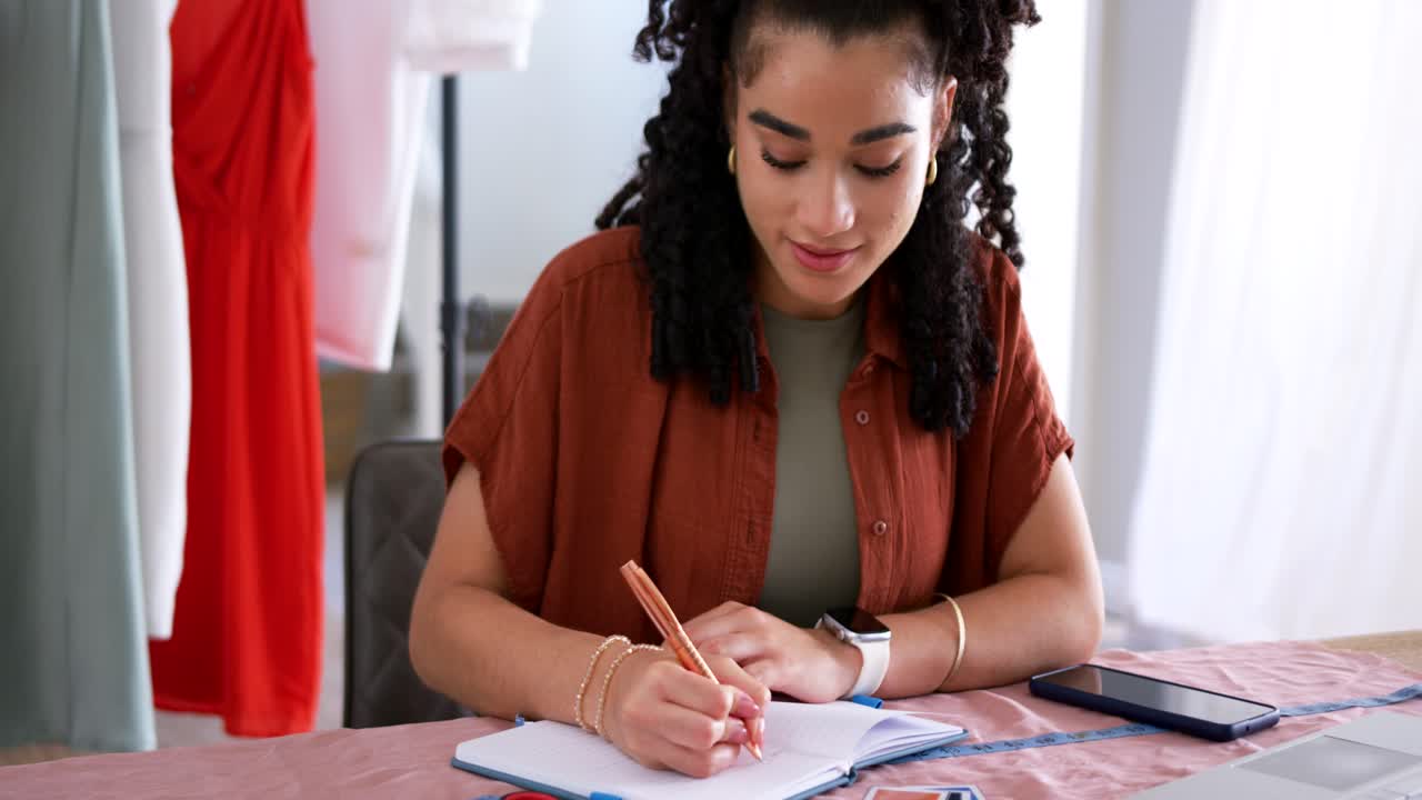 African American woman in rust top checking swatches and laptop writing notes at table designing