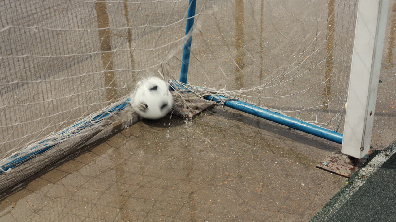 Angolo con porta bagnata e pallone da calcio che rimbalza nella rete sotto una pioggia costante, acqua che schizza alla base del palo, erba usurata e telaio in metallo blu, linea laterale vuota, impatto al rallentatore che cattura movimento e texture