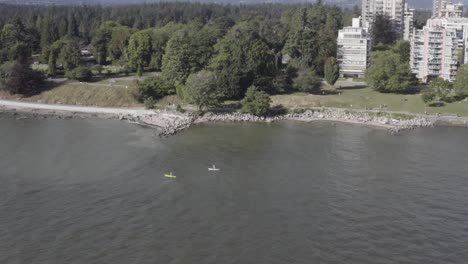 Luftfliege-180-Grad-Drehung-über-Paar-Paddle-Boarding-An-Einem-Heißen-Sonnigen-Tag-Mit-Wetsuits,-Die-An-Einem-Luxuriösen-Wohnpark-In-Der-Nähe-Des-Flachen-Endes-Des-Ozeans-Sitzen-2-2