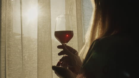 a woman with a glass of wine stands at the window at sunset