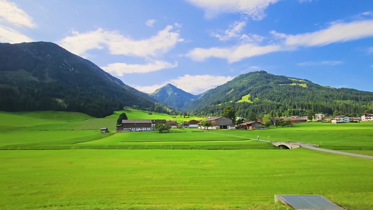 Filmato di treno in movimento che attraversa una fattoria, pendii di prati lussureggianti, colline boscose e paesaggio alpino sotto soffici nuvole estive durante un rilassante viaggio di vacanza