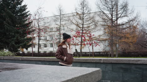 side view of lady wearing black cap brown jacket and trouser walking slowly with hands in pockets on a pathway surrounded by pine trees red-berried plants modern buildings in cold autumn atmosphere
