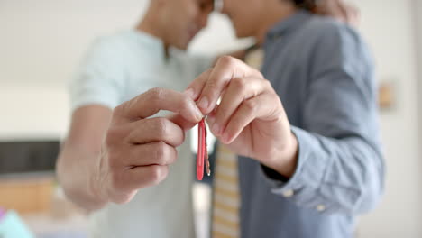 hands of diverse gay male couple holding keys to new house, embracing in living room, slow motion