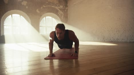 A-male-athlete-in-a-black-sports-summer-uniform-does-push-ups-on-a-special-mat-in-a-sunny-brick-hall-on-the-floor