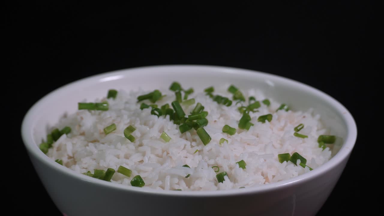 Close-up shot of a rotating bowl filled with fluffy white rice, garnished with fresh chopped chives. Set against a dark background, creating a contrast with soft lighting