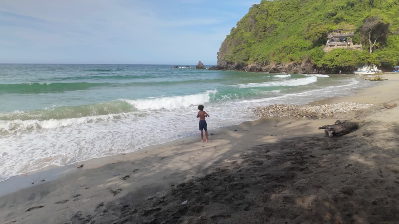 A boy fishes with nylon line on Osma Beach, Venezuela, with turquoise sea, waves, sand, and lush vegetation.