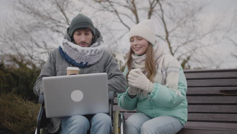 niepełnosprawny mężczyzna na wózku inwalidzkim i jego przyjaciel oglądają coś śmiesznego na laptopie i śmieją się razem w parku miejskim zimą