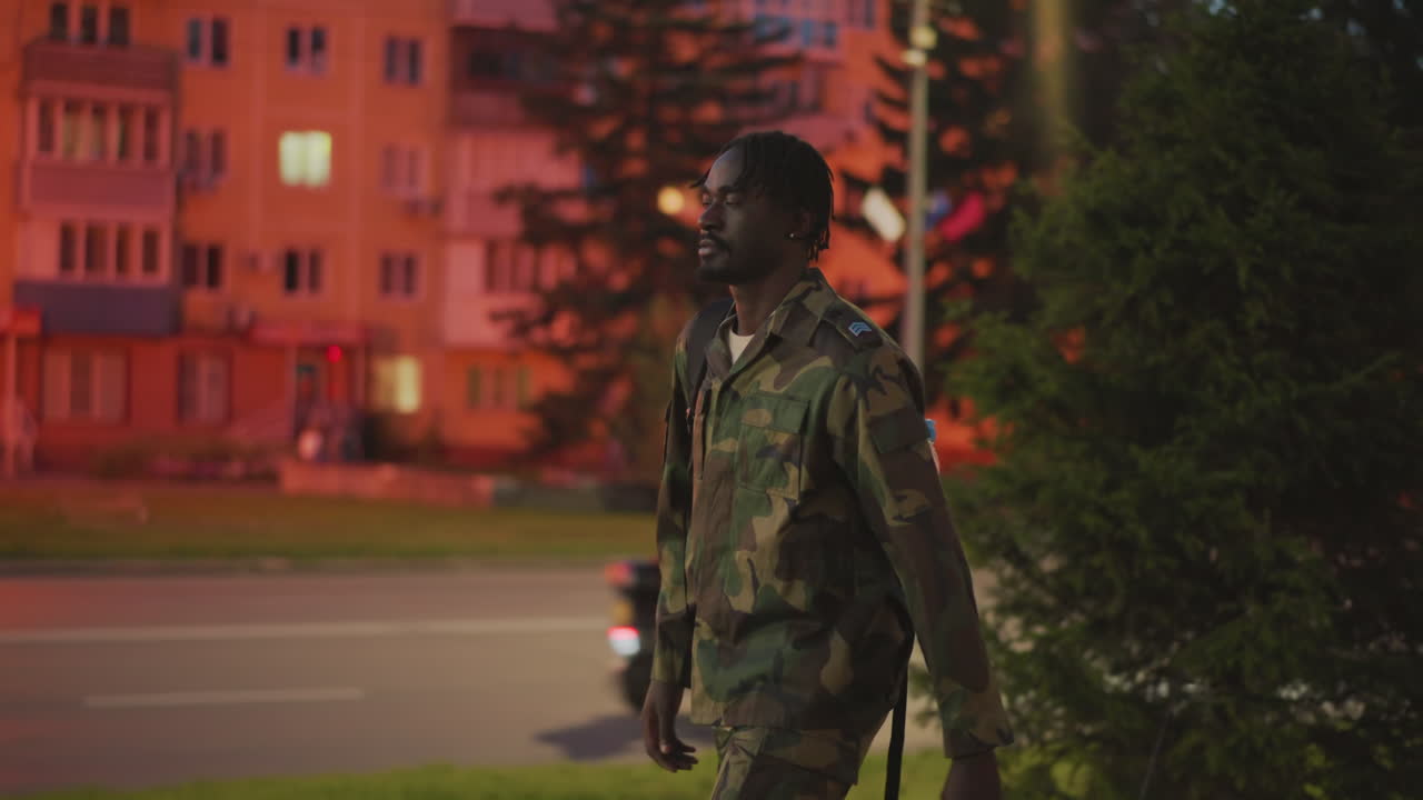 Black Soldier Patrolling Roadside Near Lit Apartment Buildings At Night, Camo Uniform And Rifle, Red Ambient Lighting From Windows, Steady Watch And Urban Tension, Solitary Guard Scene