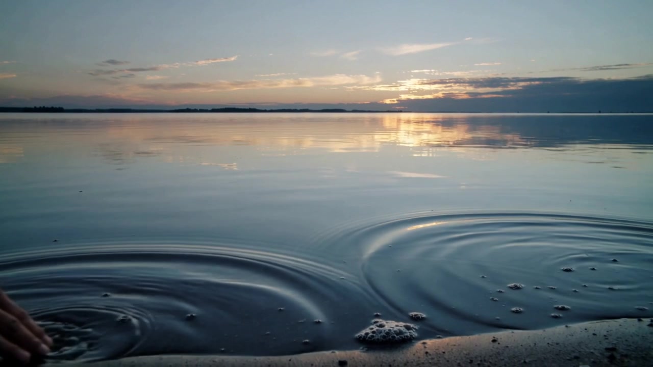 In risposta a un disturbo vicino alla riva, la superficie del lago forma anelli d'onda al tramonto, spazzando via la schiuma.