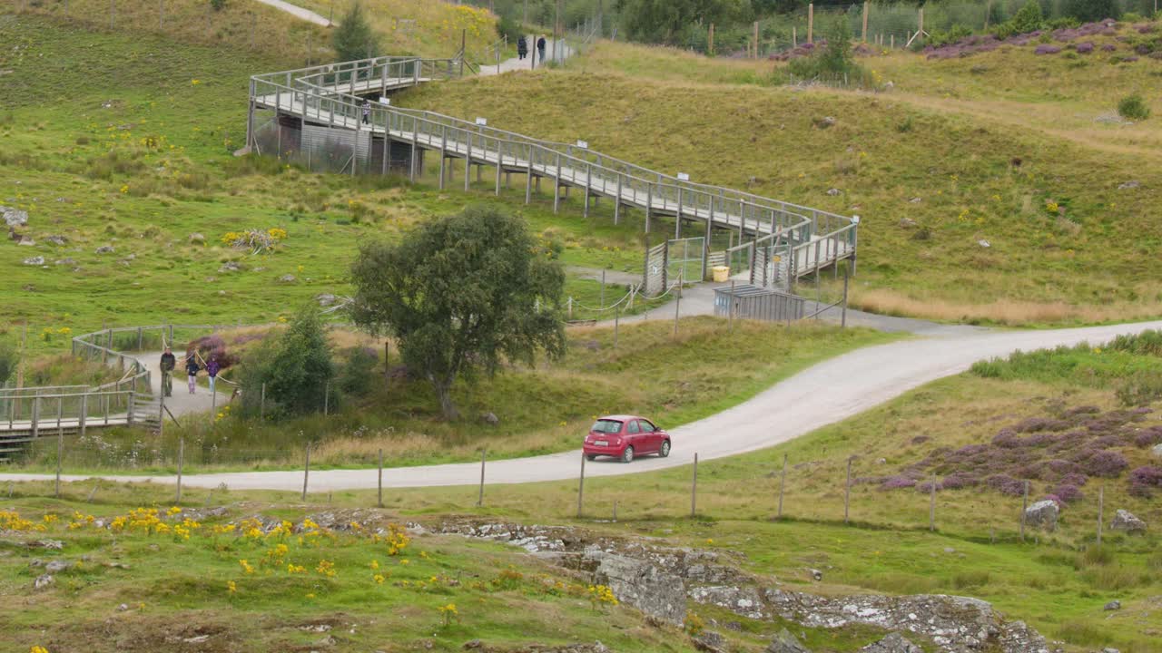 Red compact car travels along curving rural road, passing green hills and wooden pedestrian bridge