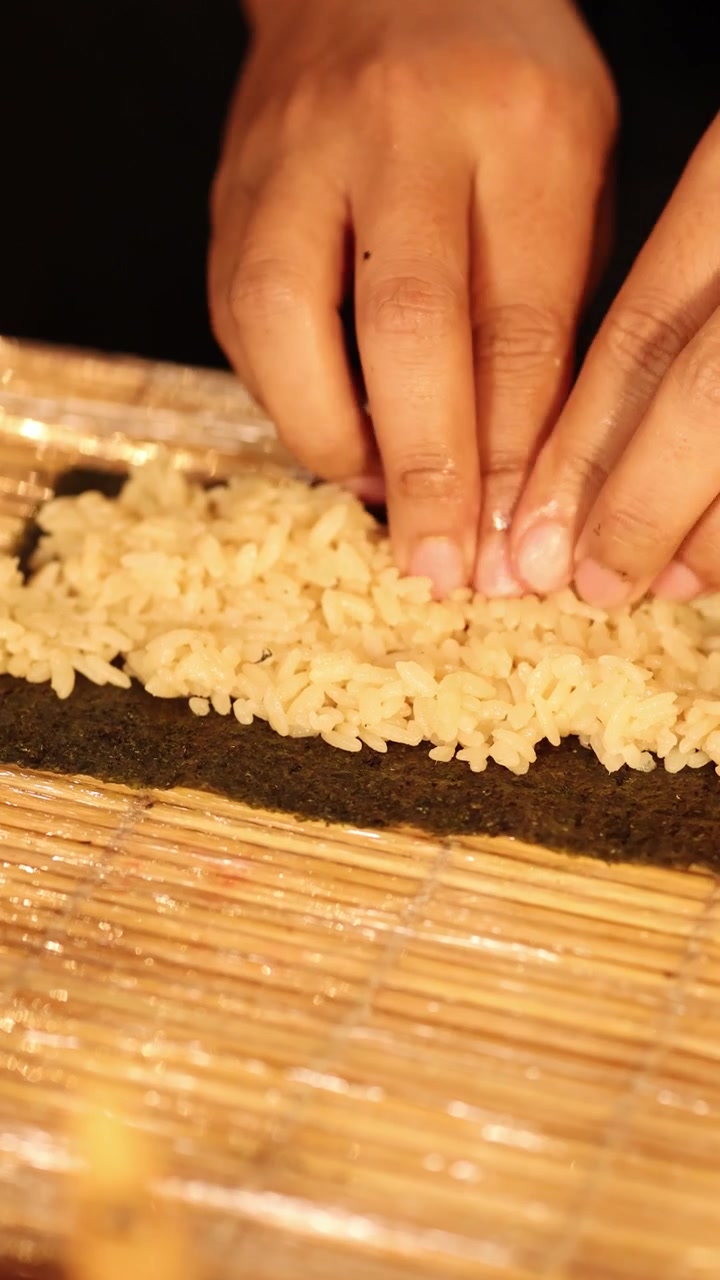 Primo piano di mani che dispongono con cura riso condito su alga nori usando una stuoia di bambù per arrotolare.