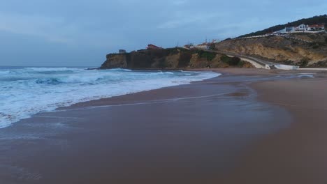 Toma-Baja-De-Drones-De-Olas-Aplastando-En-La-Playa-De-Arena-Magoito-En-Portugal,-Atardecer-Nublado