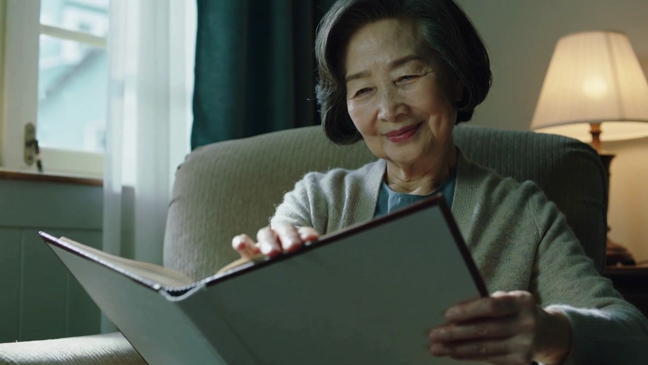 Pulling back camera showing senior Asian woman in cardigan turning pages at home, recalling album