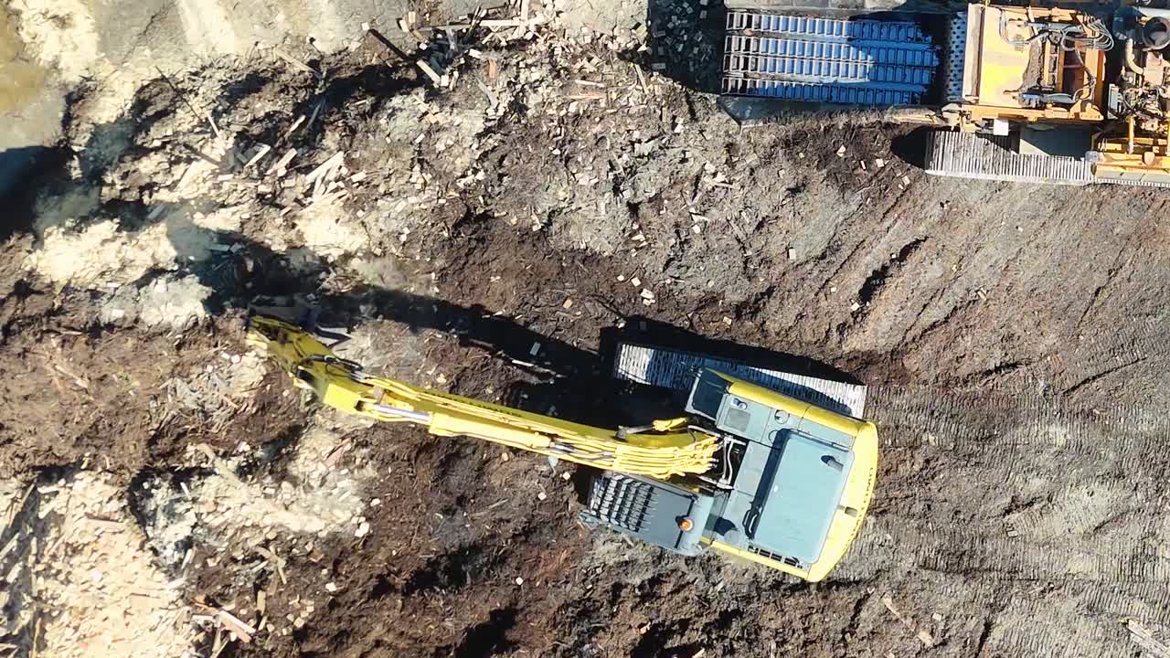 Luftfoto med drone viser en gul gravemaskine, der bearbejder mørk organisk muld på en byggeplads.