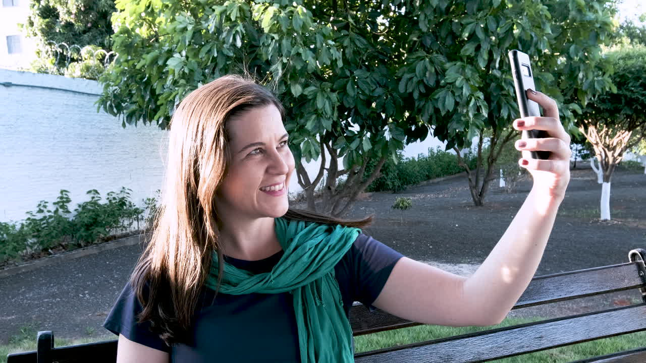 mujer joven con bufanda verde sentada en un banco en la plaza hace selfies sonrientes con árboles en el fondo y el viento