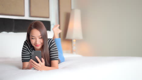 while lying on her stomach on the bed, a pretty young woman focuses on her smartphone and smiles