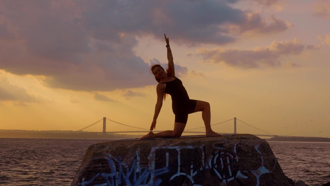 Donna in posizione yoga dell'affondo a mezzaluna con le braccia alzate al tramonto. Silhouette di profilo di una donna che si allena su un molo di pietra di fronte al ponte Verrazzano Narrows, New York