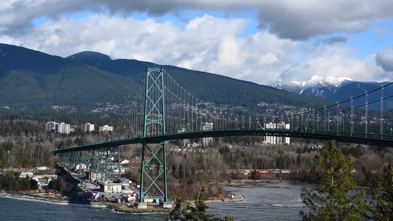 자동차가 운전하는 동안 라이온스 게이트 현수교의 파란색 기둥 아래 바위가 시간 경과에 따라 파도를 일으키고 눈 덮인 바위 산 위로 구름이 흐르고 있습니다.