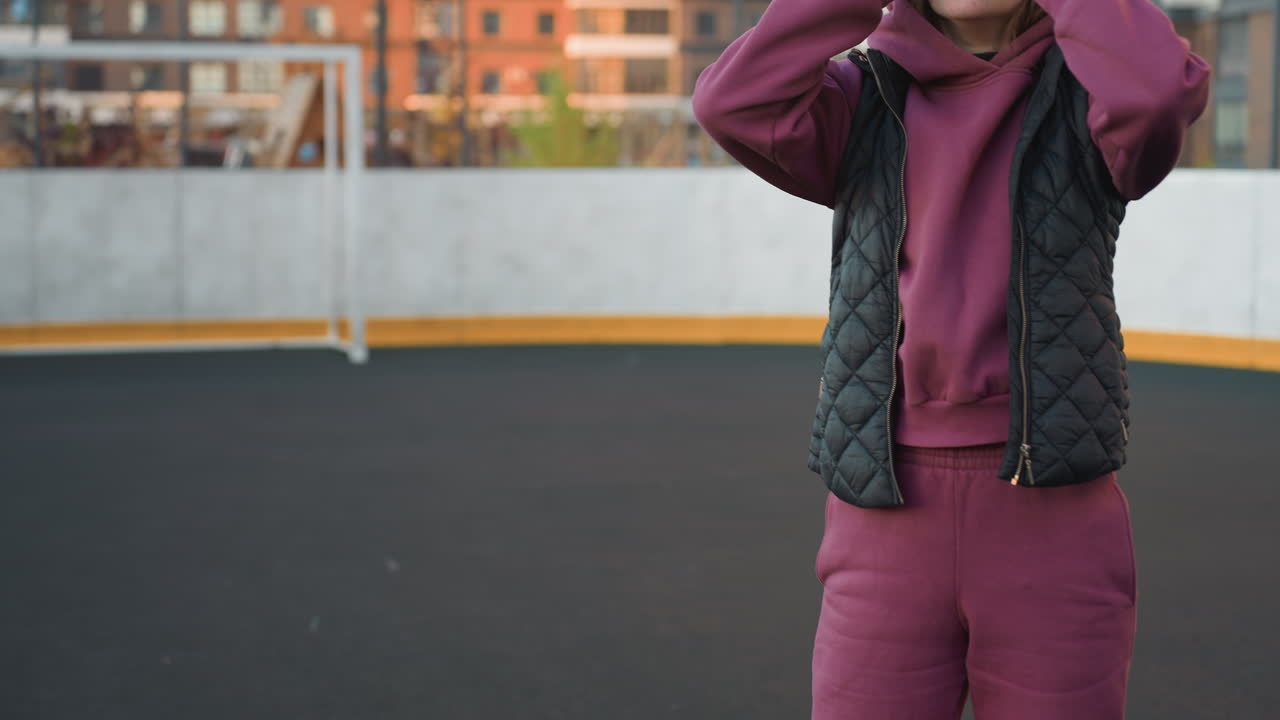 Gym goer bending down to adjust shoelaces on sneaker while wearing purple hoodie and black vest then standing up to stretch body on outdoor court under warm sunset light near tall modern buildings