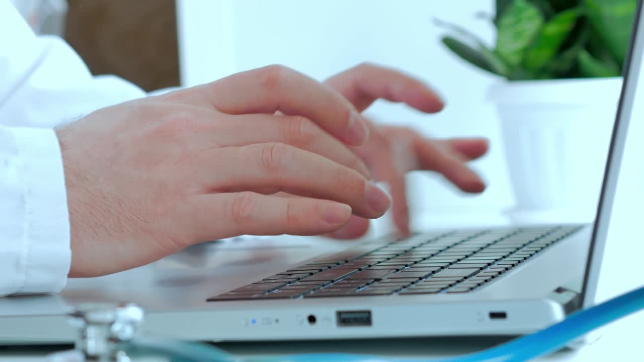 el concepto de medicina, el médico trabaja, las manos de un hombre con una túnica blanca imprimen el texto en la computadora portátil, en la mesa blanca un fanendoscopio y una planta verde dormida