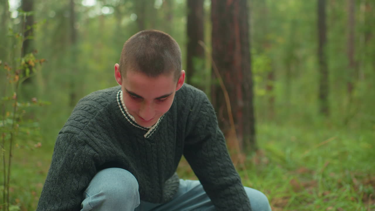 Boy in dark green knitted sweater smiling warmly as he stretches arms to set up camp tent in forest surrounded by green foliage and tall trees during outdoor adventure in peaceful woodland