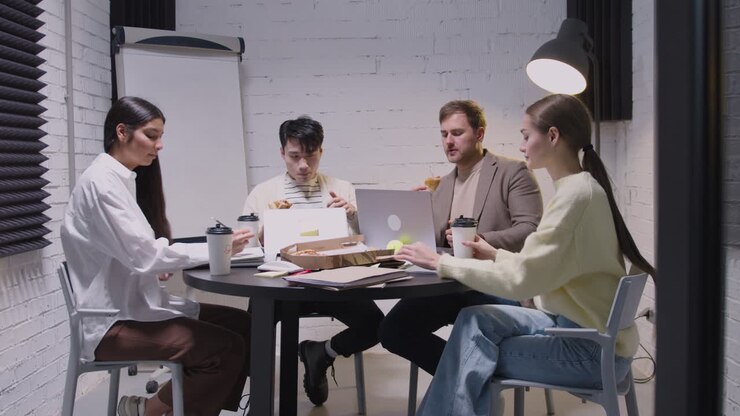 Gruppo di colleghi multietnici che mangiano pizza e bevono caffè da asporto durante una riunione di squadra nella sala riunioni