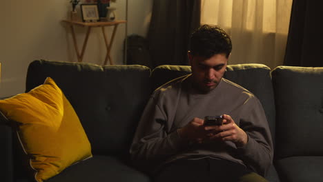 young man spending evening at home sitting on sofa with mobile phone scrolling through internet or social media 4