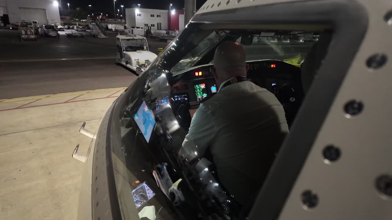 An external view through the cockpit window, of a pilot doing the pre-flight checks before the first flight of the day at night, with a side view of the cockpit of a modern jet