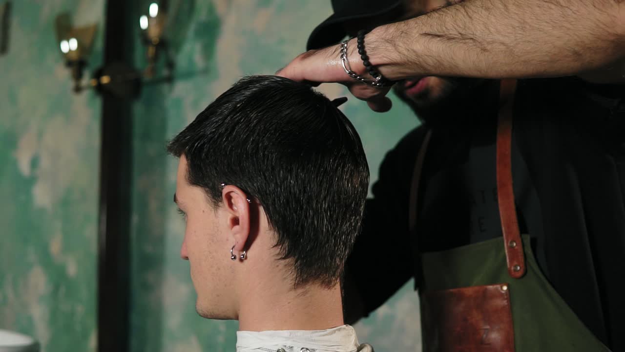 un peluquero peina suavemente el cabello corto y húmedo del cliente masculino. barbero barbudo en delantal peinando el cabello de un cliente masculino sentado en el