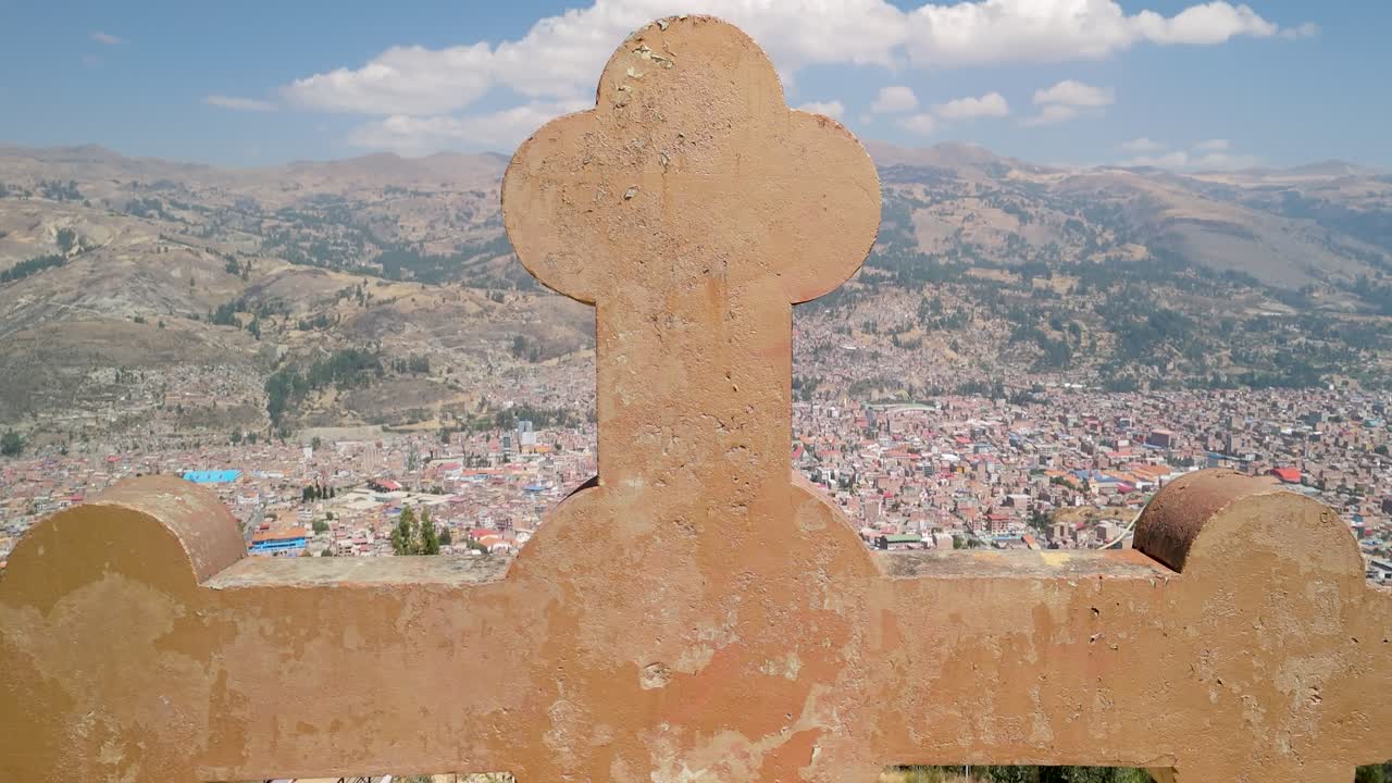 An aerial shot rises and tilts down, revealing the sprawling Huaraz cityscape behind the large stone cross at the famous Rataquenua viewpoint in the Peruvian Andes