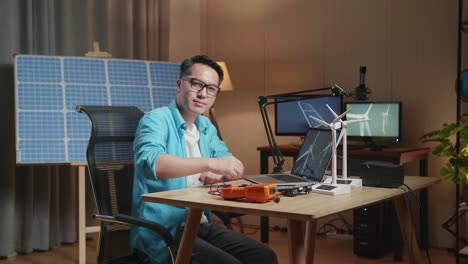 hombre asiático mira la turbina eólica, cruza los brazos y sonríe a la cámara mientras trabaja con una computadora portátil junto a la célula solar en la oficina