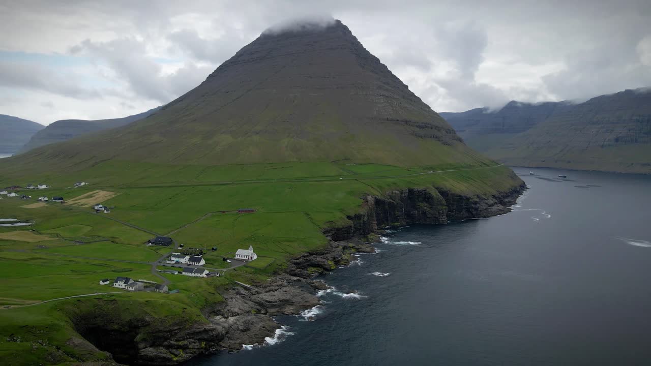 una escarpada montaña se eleva sobre el sereno pueblo de viðareiði en las islas feroe, rodeada de exuberante vegetación, acantilados escarpados y vistas al océano. una escena tranquila que mezcla naturaleza y cultura