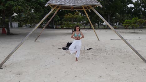 Kaukasische-Brünette-Junge-Dame-Praktiziert-Yoga-Mit-Weißen-Hosen-Am-Strand-In-Asien-Luftdrohne-Panoramablick-In-Hölzernen-Pavillon