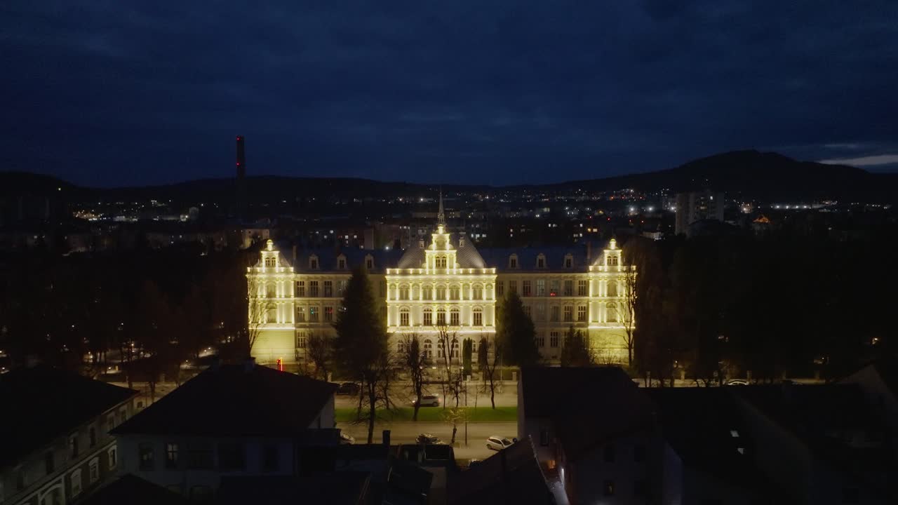 Wide aerial shot, smooth push-out movement revealing the historic College National "Liviu Rebreanu" in Bistrita Romania. Beautifully illuminated against the blue night sky with city lights background