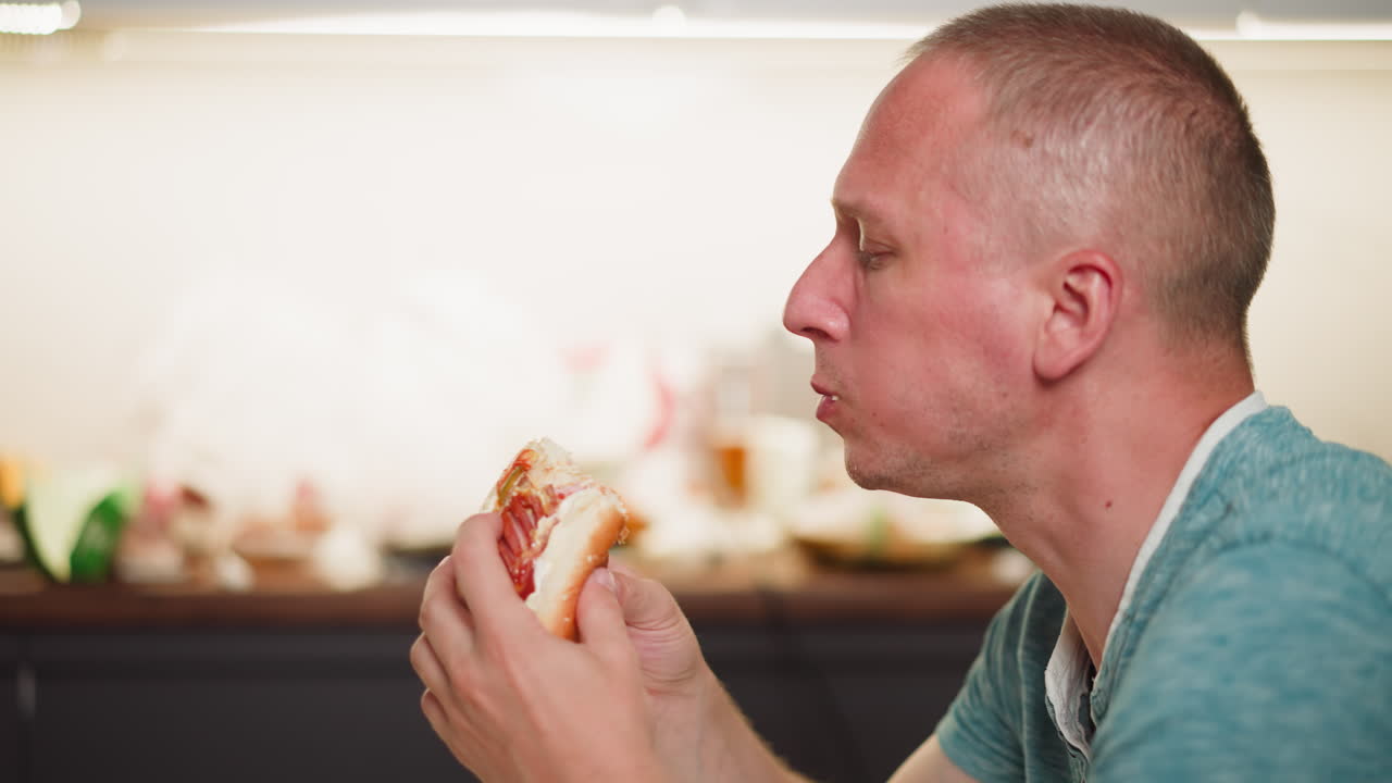 Side view of man eating sausage sandwich indoors, holding bread filled with toppings in hands, cheeks puffed while chewing, wearing casual teal shirt with blurred kitchen background